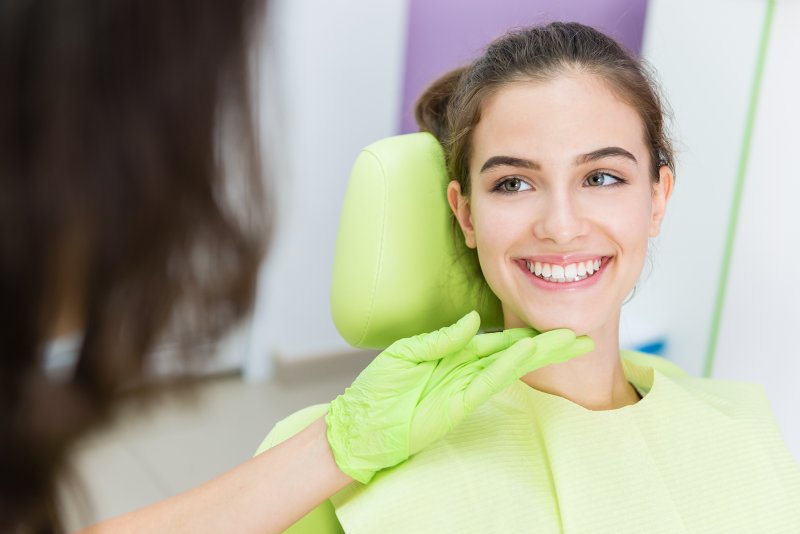 Girl sitting in dental chair smiling with teeth as dentist holds gloved hand up to her chin